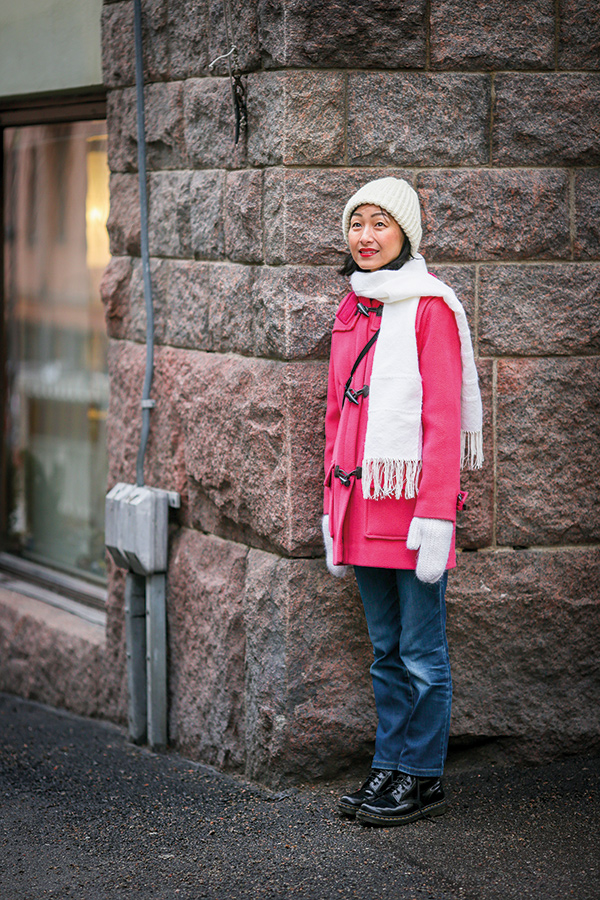 Hisayo Katsui, en japansk kvinna med ljusröd rock och vit scarf lutar sig mot en stenvägg.