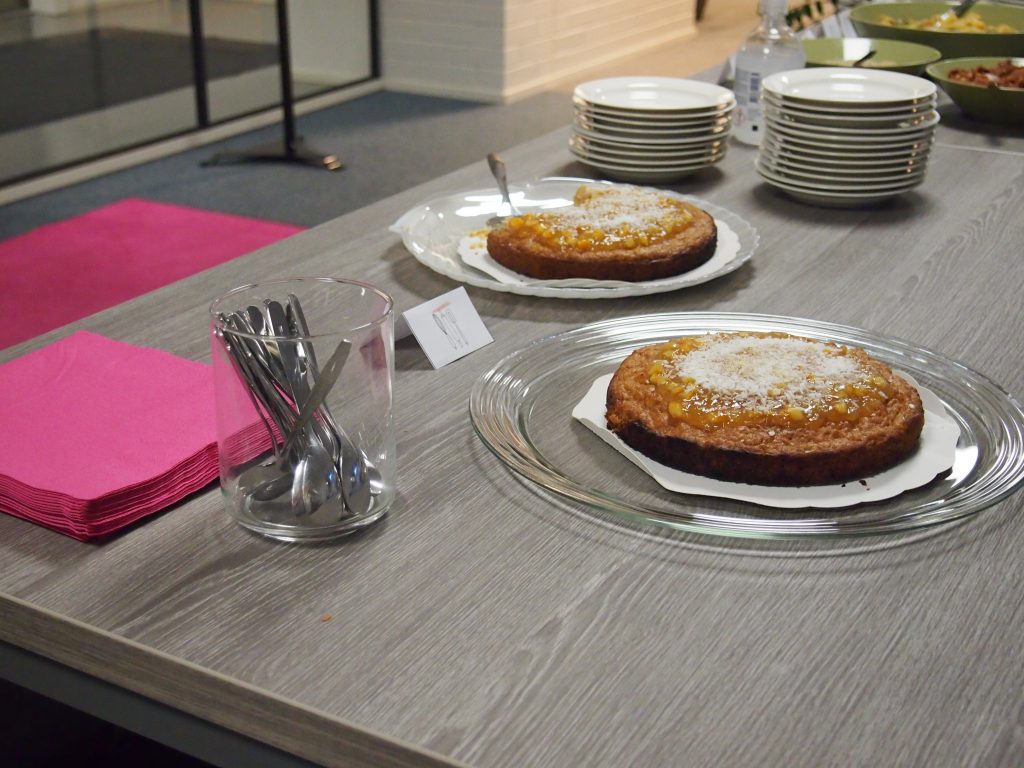 Två kakor på serveringsfat på ett bord. Rosa servetter och teskedar i ett glas. Två staplar med vita fat.