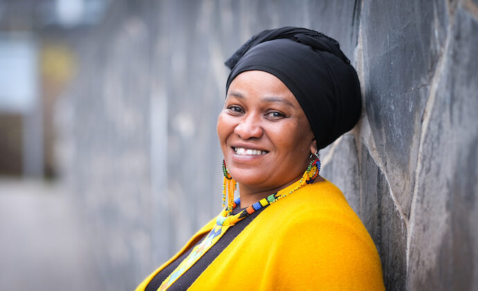 Porträttbild på Fatma Wangare. Hon har en gul klädsel, en svart skarf på huvudet och färggranna smycken. Hon ler brett mot kameran.