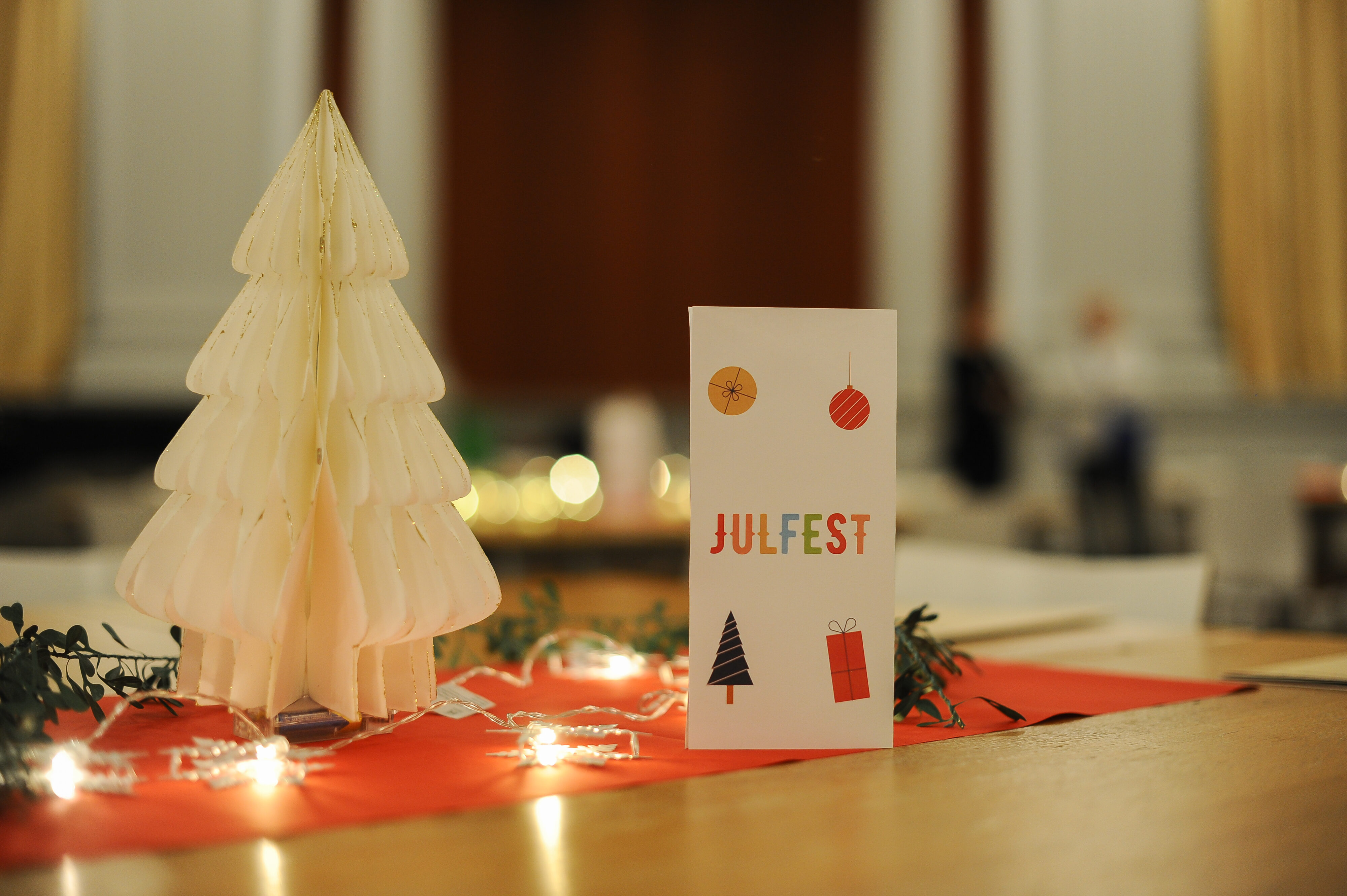 Ett bord fotat på nära håll, med en vit pappersgran och en broschyr med texten Julfest. En röd bordduk, en lysande ljusslinga och en grön girland.