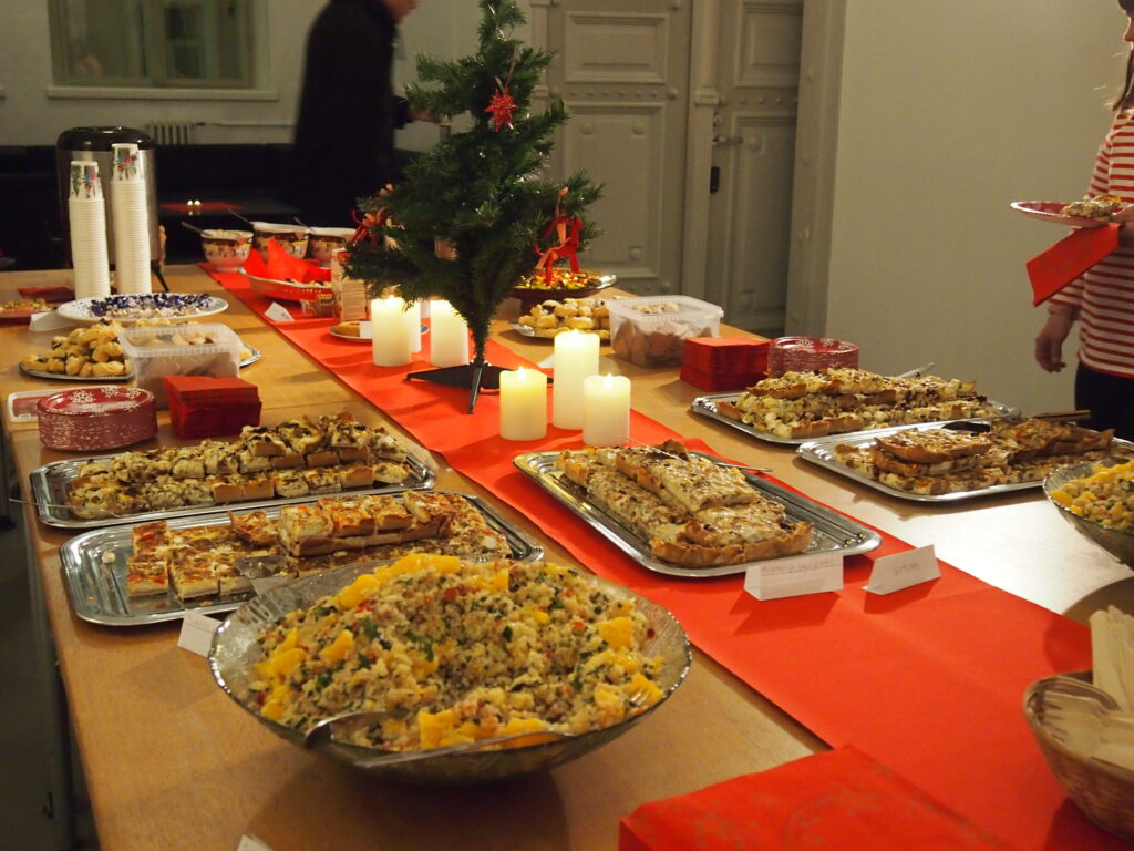 Ett bord med röd bordduk och liten julgran. Olika former av mat såsom sallad, pajbitar och jultårtor. 
