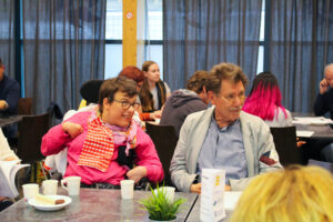 Två deltagare sitter vid ett bord, andra deltagare syns i bakgrundne. På bordet finns kaffekoppar.