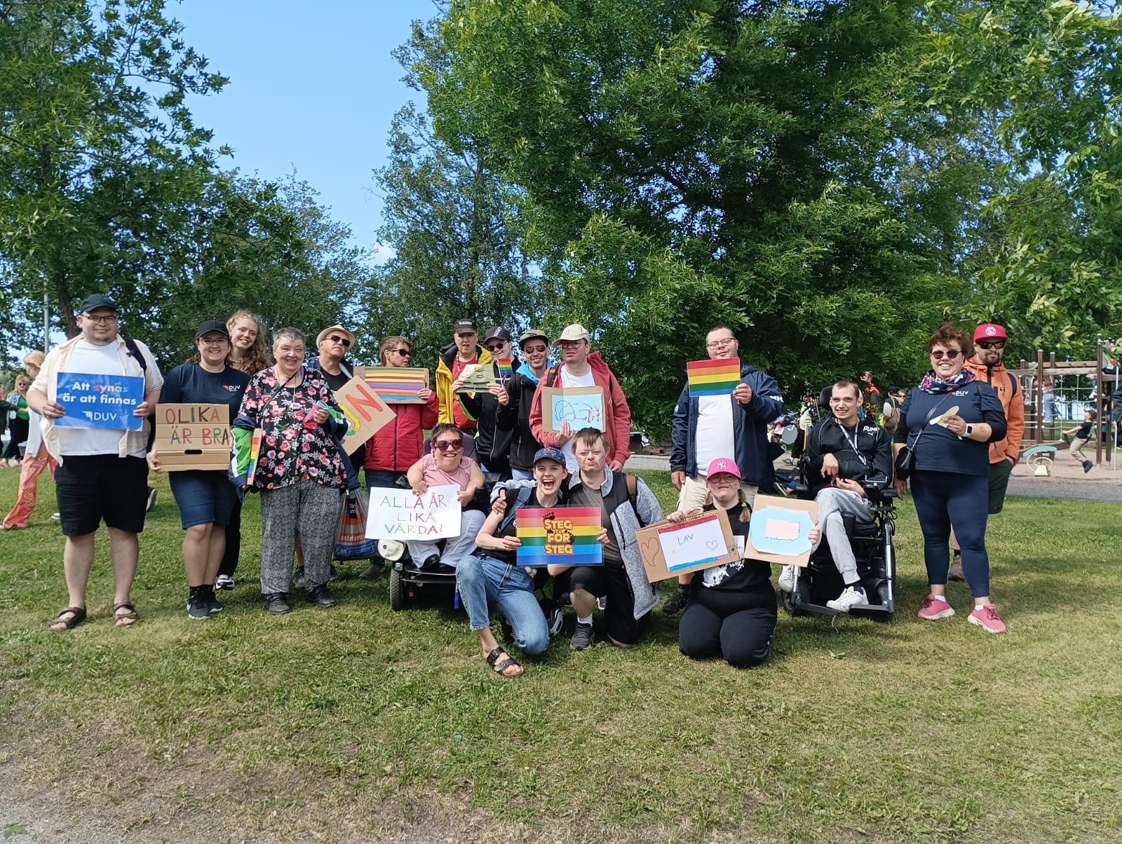 Deltagare på Vasa pride 2024. Personerna står i en park och håller i olika plakat och prideflaggor.