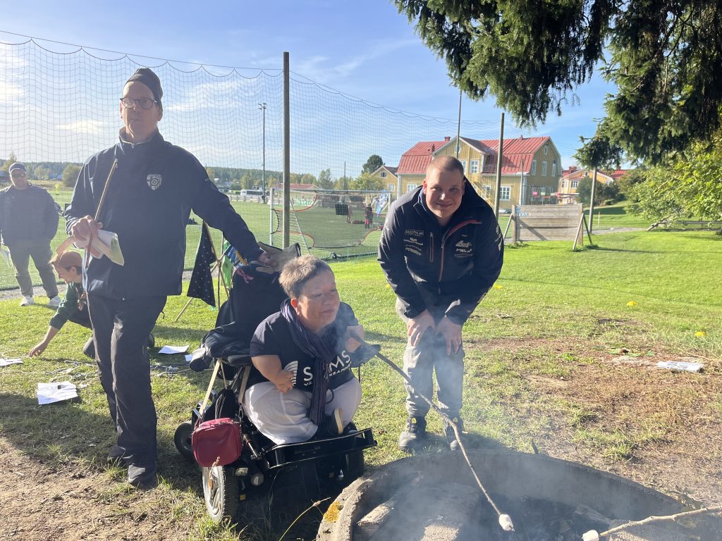 Tre personer står framför en grillplats varav en person grillar en marshmallow.