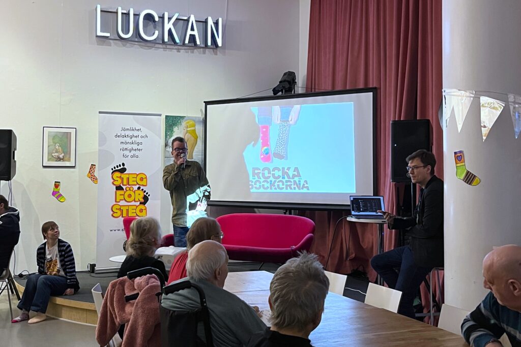 Steg för stegs ordförande Jon står på scen och hälsar Rocka sockorna-deltagarna välkomna. I förgrunden syns deltagare sittande vid bord.