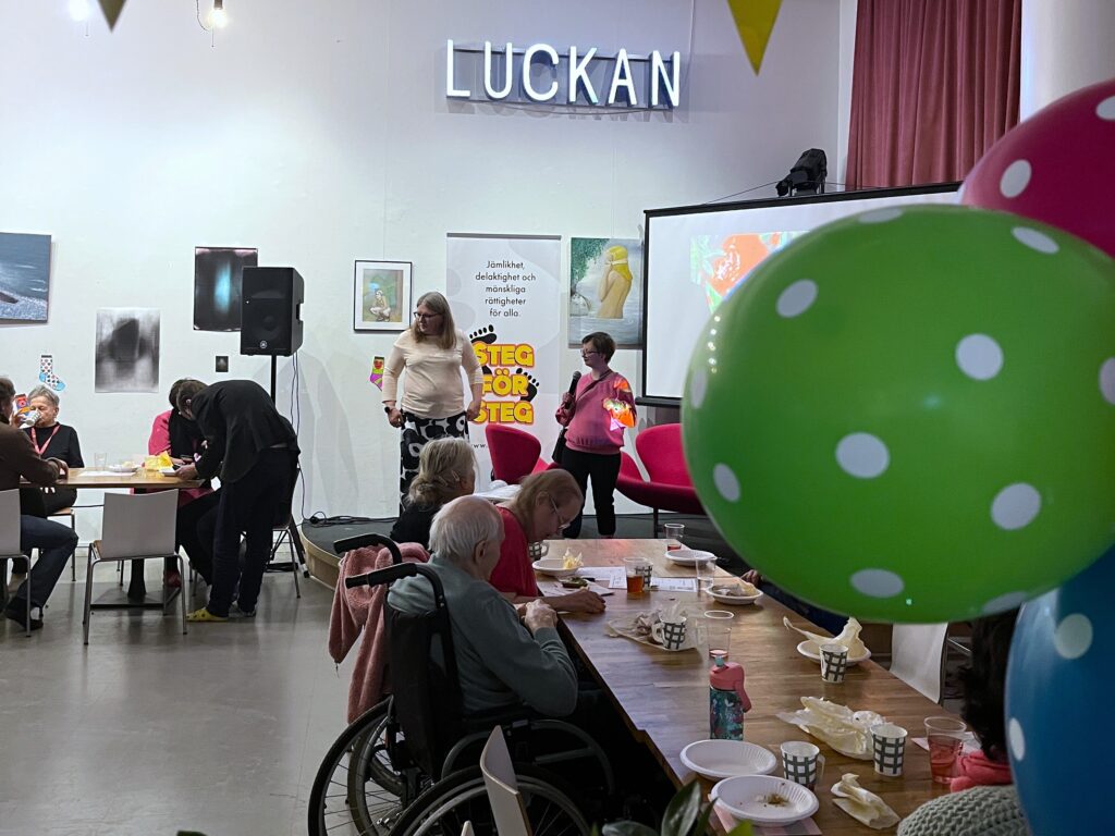 Två personer sjunger på scen under programpunkten talangshow på Rocka sockorna-festen; i förgrunden sitter deltagare vid bord och en stor prickig ballong syns till höger.